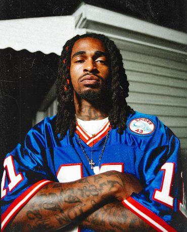 Man with dreadlocks wearing a blue sports jersey poses confidently with crossed arms.