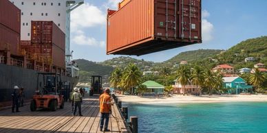 A shipping container is lifted at a tropical dock with workers around.