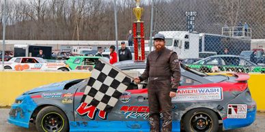 Race car driver stands next to winning car with trophy and checkered flag.
