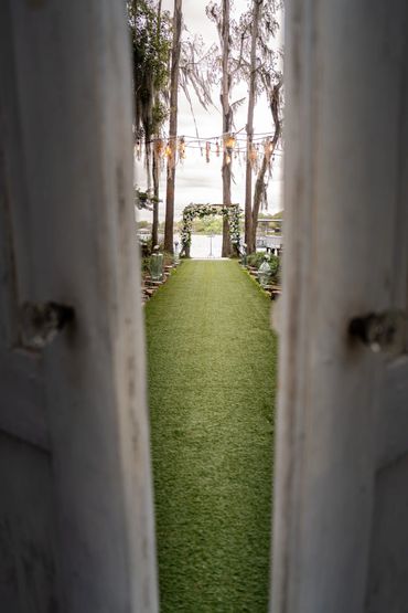 View through a doorway of a green aisle leading to a floral wedding arch outdoors.