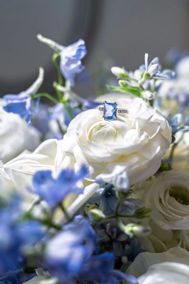 Blue gemstone ring on a white rose surrounded by blue flowers.