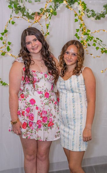 Two young women smiling, dressed in floral and patterned summer dresses, standing in front of a decorated backdrop.