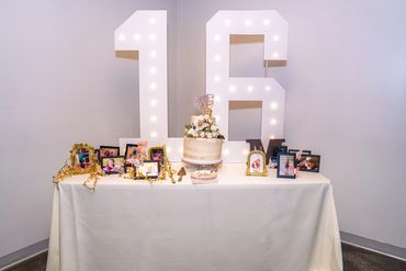 Elegant 16th birthday cake table with illuminated number lights and framed photos.