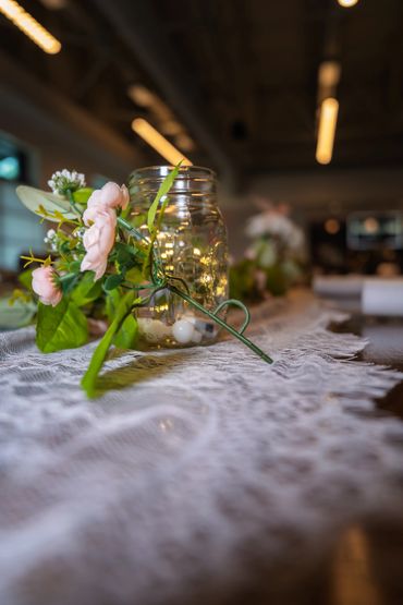 A mason jar with fairy lights and flowers on a lace table runner.