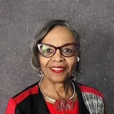 Smiling elderly woman wearing glasses and a red outfit with a leaf necklace.