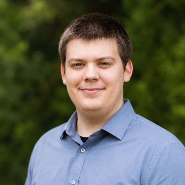 Smiling man in a blue shirt outdoors with green foliage background.
