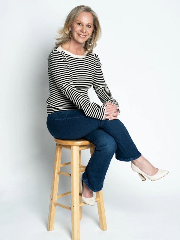 Smiling woman in striped top sitting on wooden stool against white background.