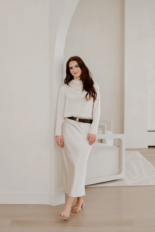 A woman in a cream dress stands gracefully indoors.
