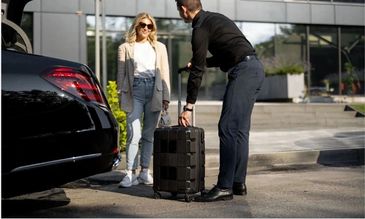 A man helps a woman with her suitcase by a car outside a modern building.