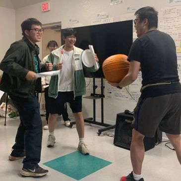 Four people engaging in a playful indoor activity with a pumpkin and paper plates.