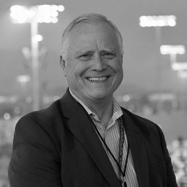 Smiling elderly man in a blue suit outdoors at dusk.