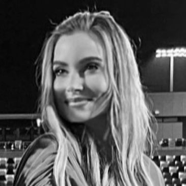 A woman in a blue jacket smiles on a lit soccer field at night.