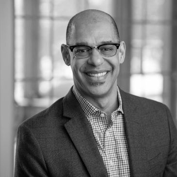 Smiling man wearing glasses and a blazer in a softly lit room.