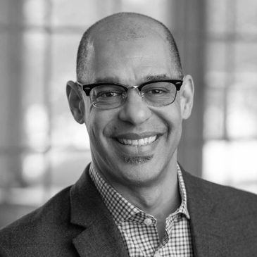 Smiling man wearing glasses and a blazer in a softly lit room.