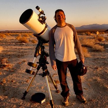Man standing next to a telescope in a desert during sunset.