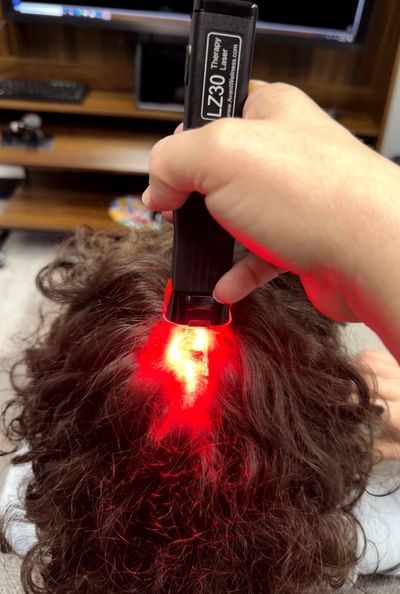 Hand holding a red light therapy device over curly hair.