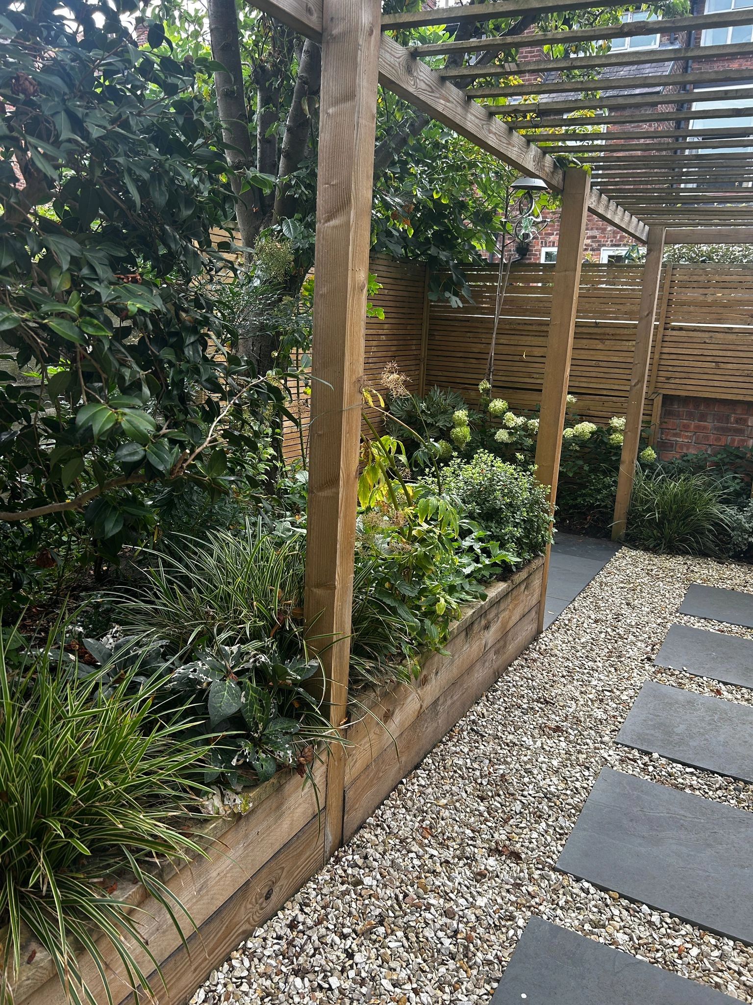 A wooden pergola over a garden bed with greenery and a stone pathway of large square slabs.