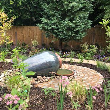 A garden with a large tipped ceramic pot water feature surrounded by plants.