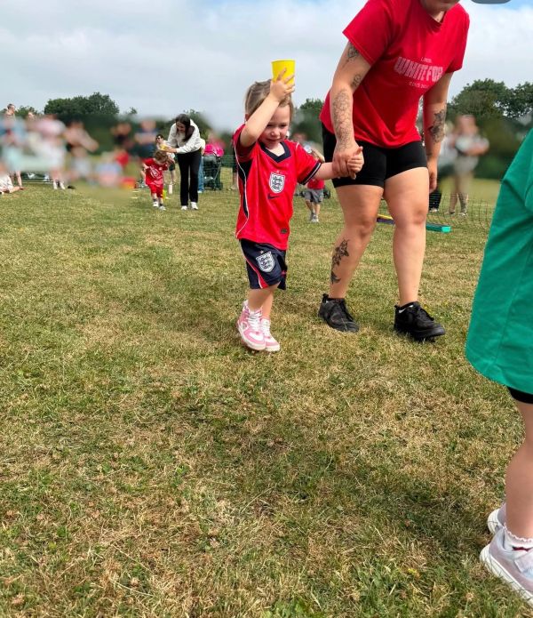 Child holding a yellow cup, holding hands with an adult in a sports field.