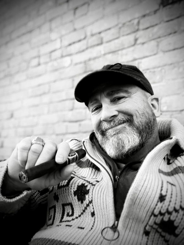 Man in a cap and patterned sweater holding a cigar, smiling.