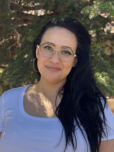 Woman with glasses and long dark hair smiling outdoors.