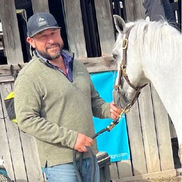 Man in green sweater holding a white horse by the reins outside a wooden stable.