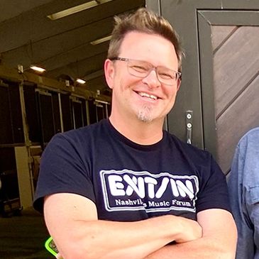 Smiling man in glasses and a music forum t-shirt stands with arms crossed.