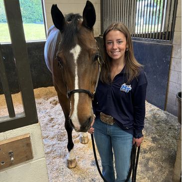 Young woman standing beside a brown horse in a stable, smiling.
