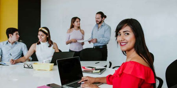 A woman in red works on a laptop while colleagues discuss in the background.