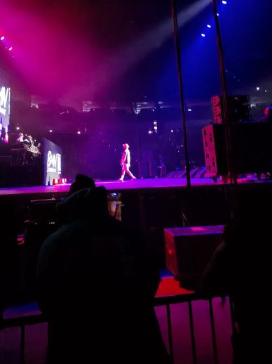Performer walking on stage under vibrant purple and blue lights during a concert.