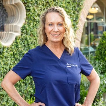 Smiling woman in blue medical scrubs posing outdoors with greenery background.