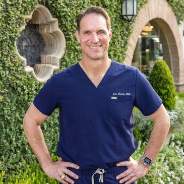 Smiling doctor in navy scrubs standing outdoors with hands on hips.