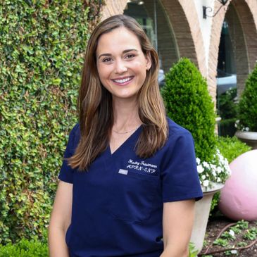 Smiling woman in navy medical scrubs outdoors near greenery and arches.
