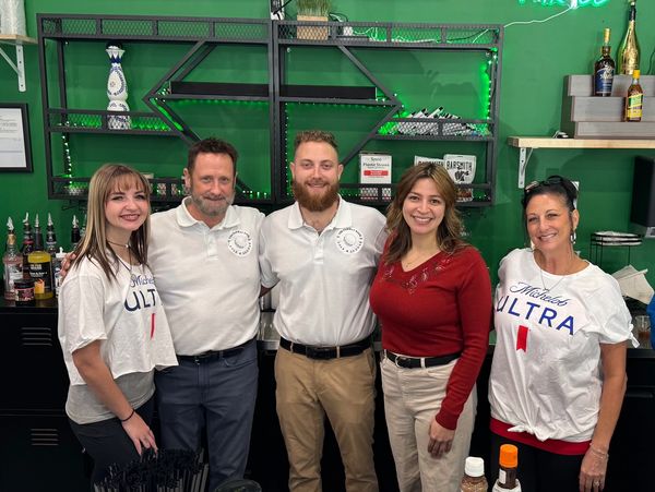 Group of five people smiling behind a bar with green walls and neon sign.