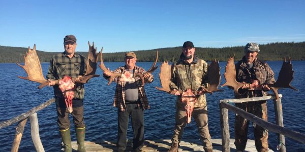 Four men holding moose antlers by a lake.