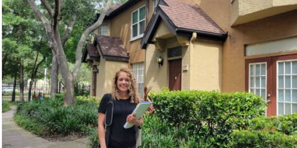A woman with curly hair stands outside a residential building holding papers and smiling.