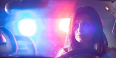 A woman in a car looks concerned with police lights flashing behind her.