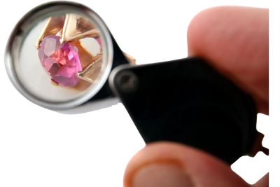 A pink gemstone viewed through a jeweler's loupe held by a hand.