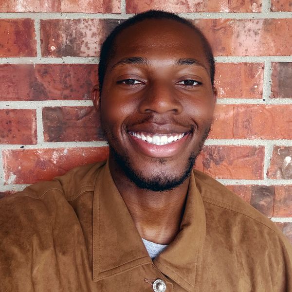 Smiling man in brown jacket poses against a brick wall.