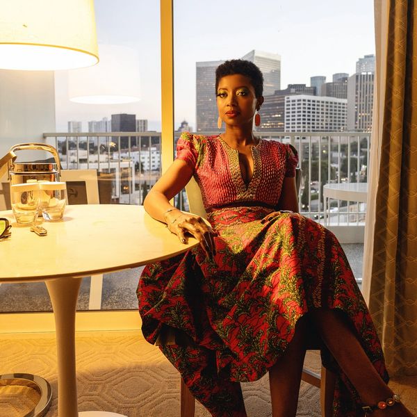 Woman in a vibrant patterned dress sitting indoors by a table with cityscape background.