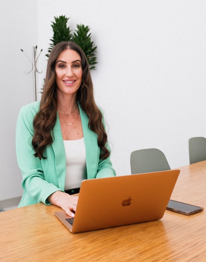 Professional woman in a mint blazer working on a laptop at a wooden table.