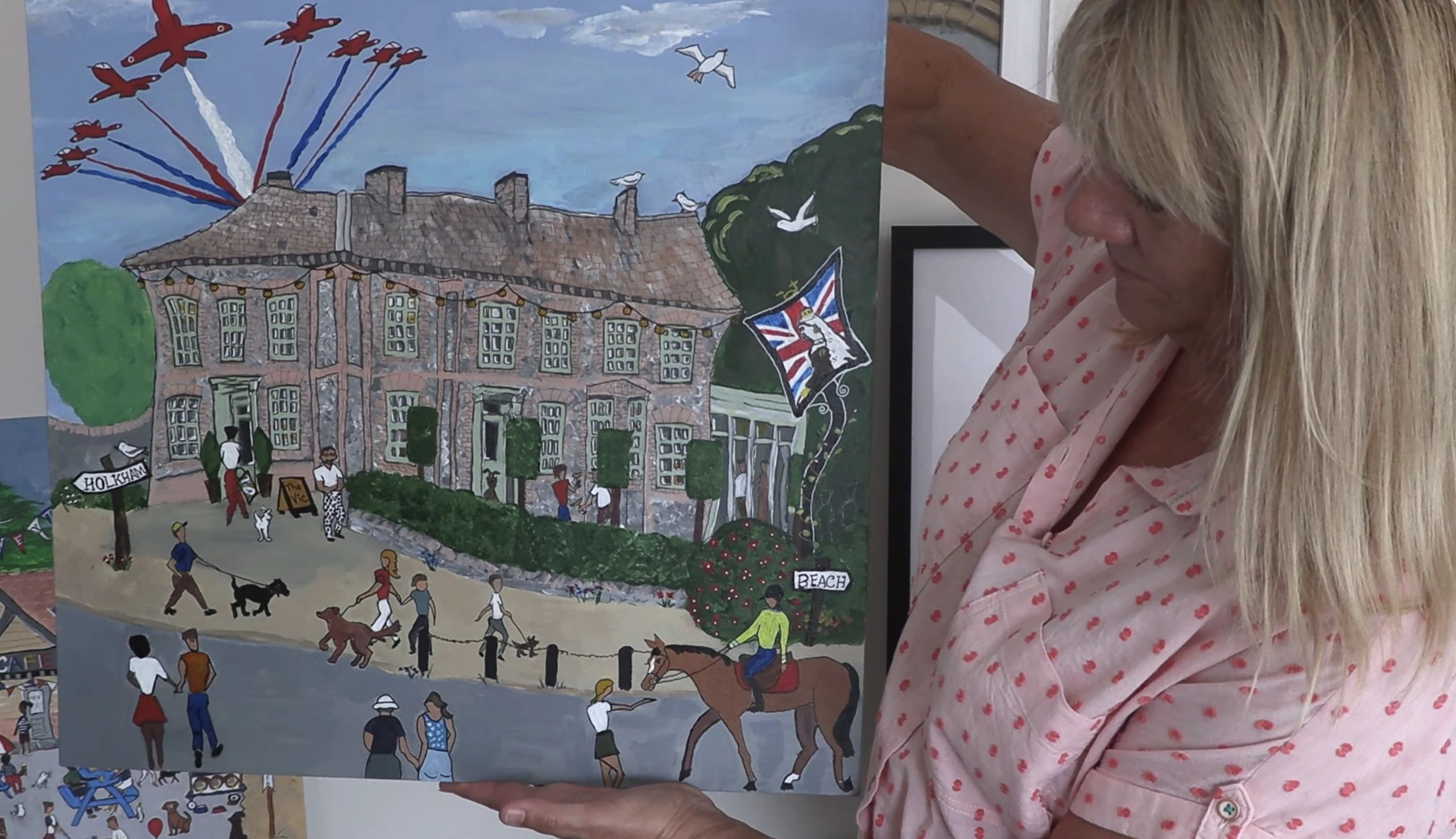 A woman holds a colorful painting featuring a large house, people, dogs, and planes in the sky.
