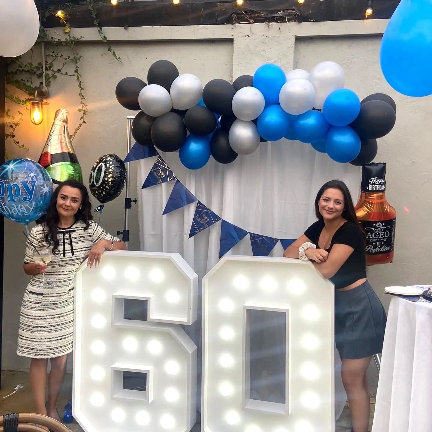 Two women celebrating a 60th birthday with balloons and lit numbers.