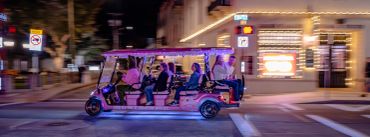 People riding a pink-lit golf cart at night in a city.