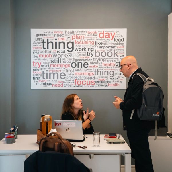 Two colleagues discuss work at a desk with a motivational word cloud poster behind them.