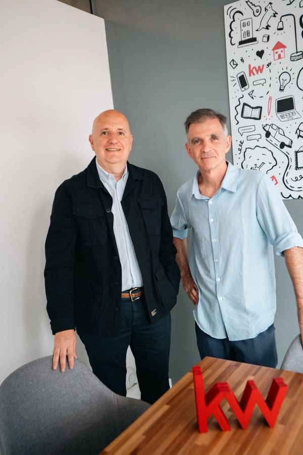 Two men standing beside a table with a red "kw" logo sculpture.