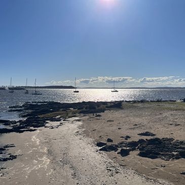Sailboats anchored on a sunny, shimmering bay with a rocky shoreline.
