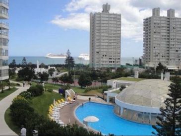Coastal cityscape with pool, tall buildings, and cruise ships at sea.