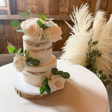 Two-tier cake adorned with white roses and greenery on a rustic wooden base.
