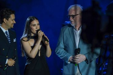 Three people on stage, two men and a woman singing or speaking.